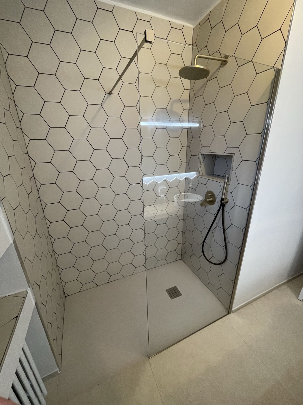 Douche à l'italienne en carrelage hexagonal blanc avec robinetterie dorée et niche de rangement, à la place de l'ancienne baignoire